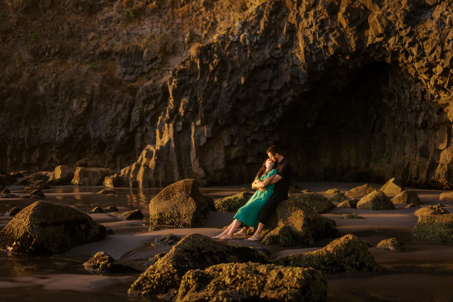 Couple embracing on mossy rocks inside a dramatic sea cave at golden hour on a scenic coastal beach.