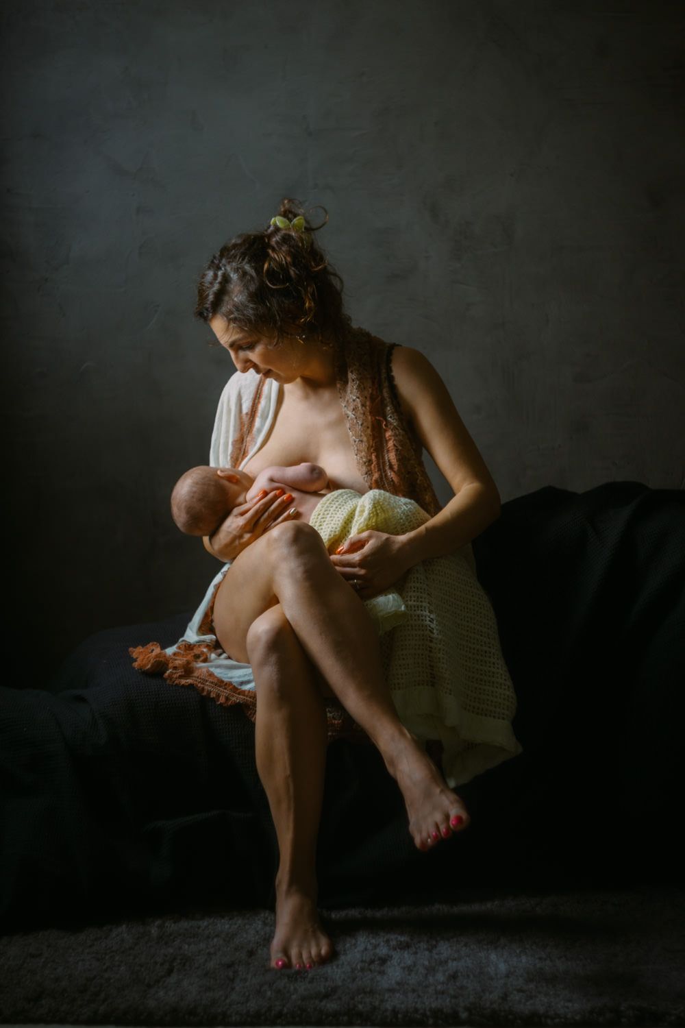 A mother tenderly breastfeeds her newborn baby while seated on a dark sofa in a softly lit room.