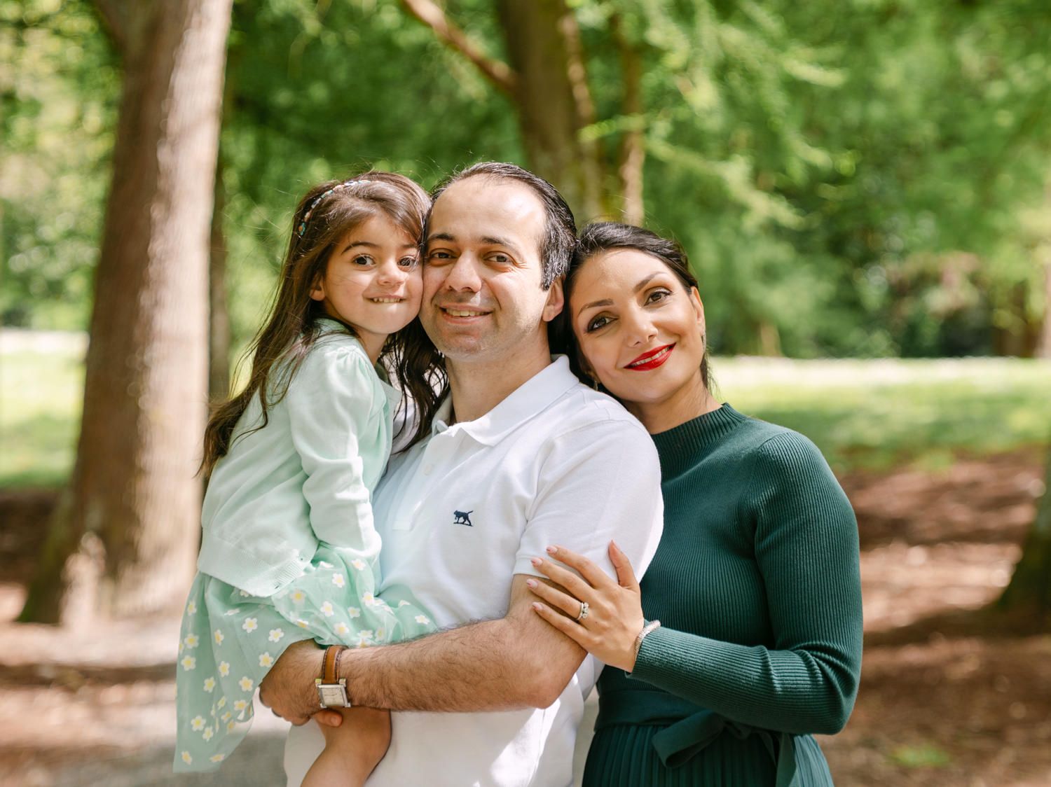 Happy family of three posing outdoors in a lush green park, parents holding young daughter.