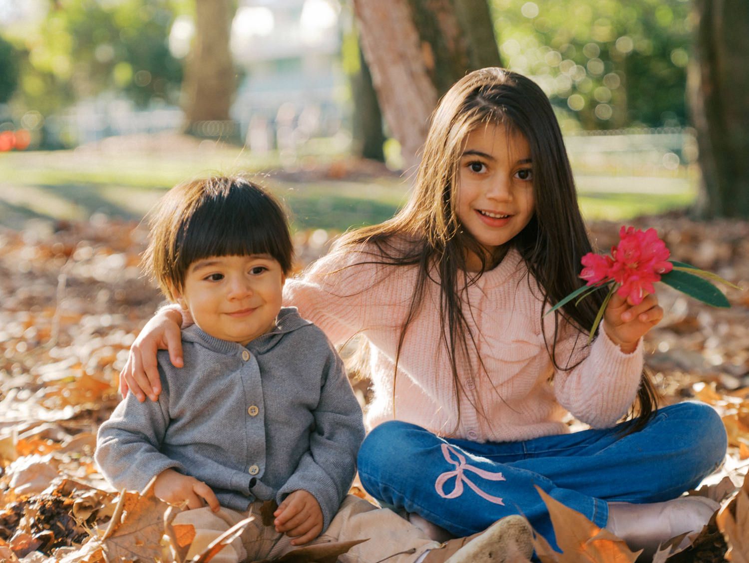 Two young siblings sit among autumn leaves in a park, with a girl holding a pink flower smiling beside her toddler brother.