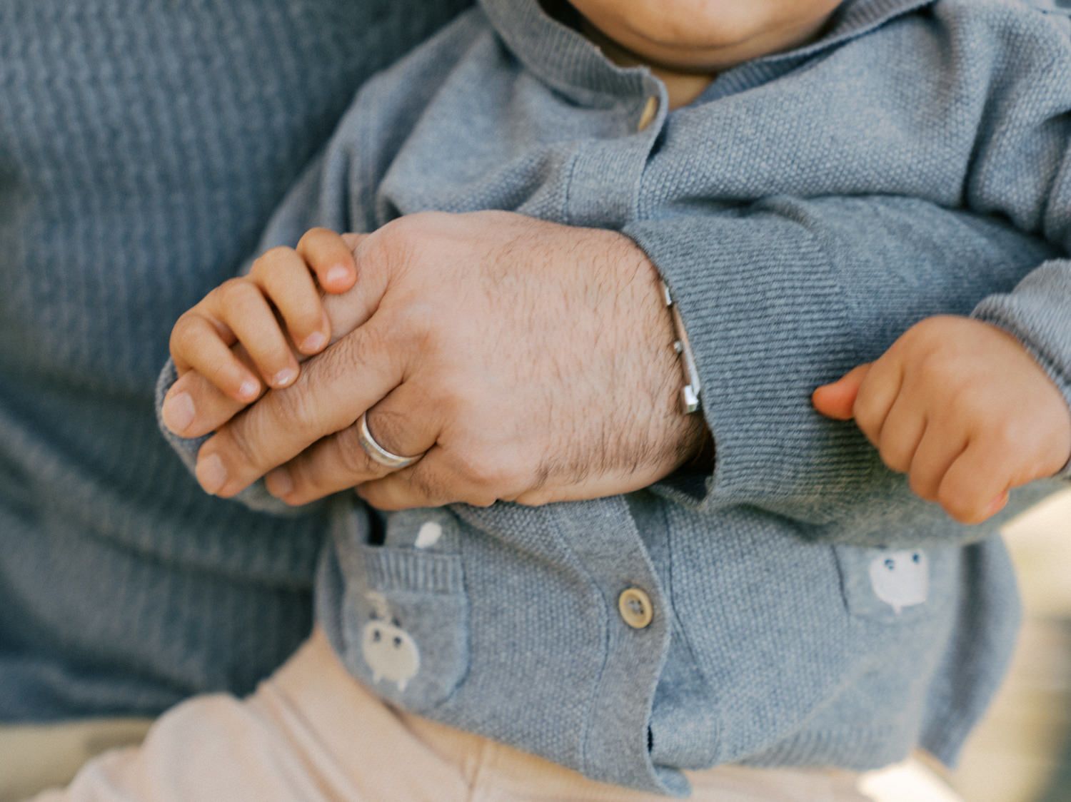 Adult hand with bracelet and ring gently holding a baby dressed in a gray knit sweater with bear details.