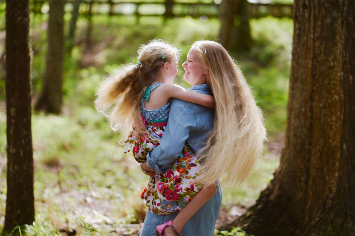 A mother with long blonde hair holds her young daughter in a sunlit forest, both smiling and sharing a tender moment.
