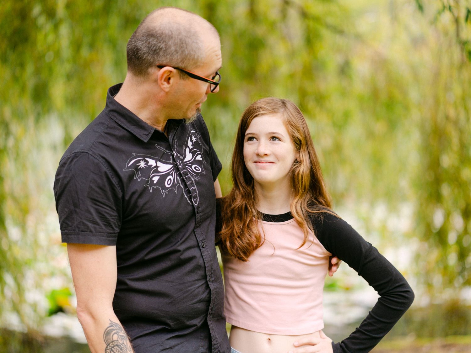 A bald man and teenage girl with red hair share a warm moment outdoors near weeping willow trees.