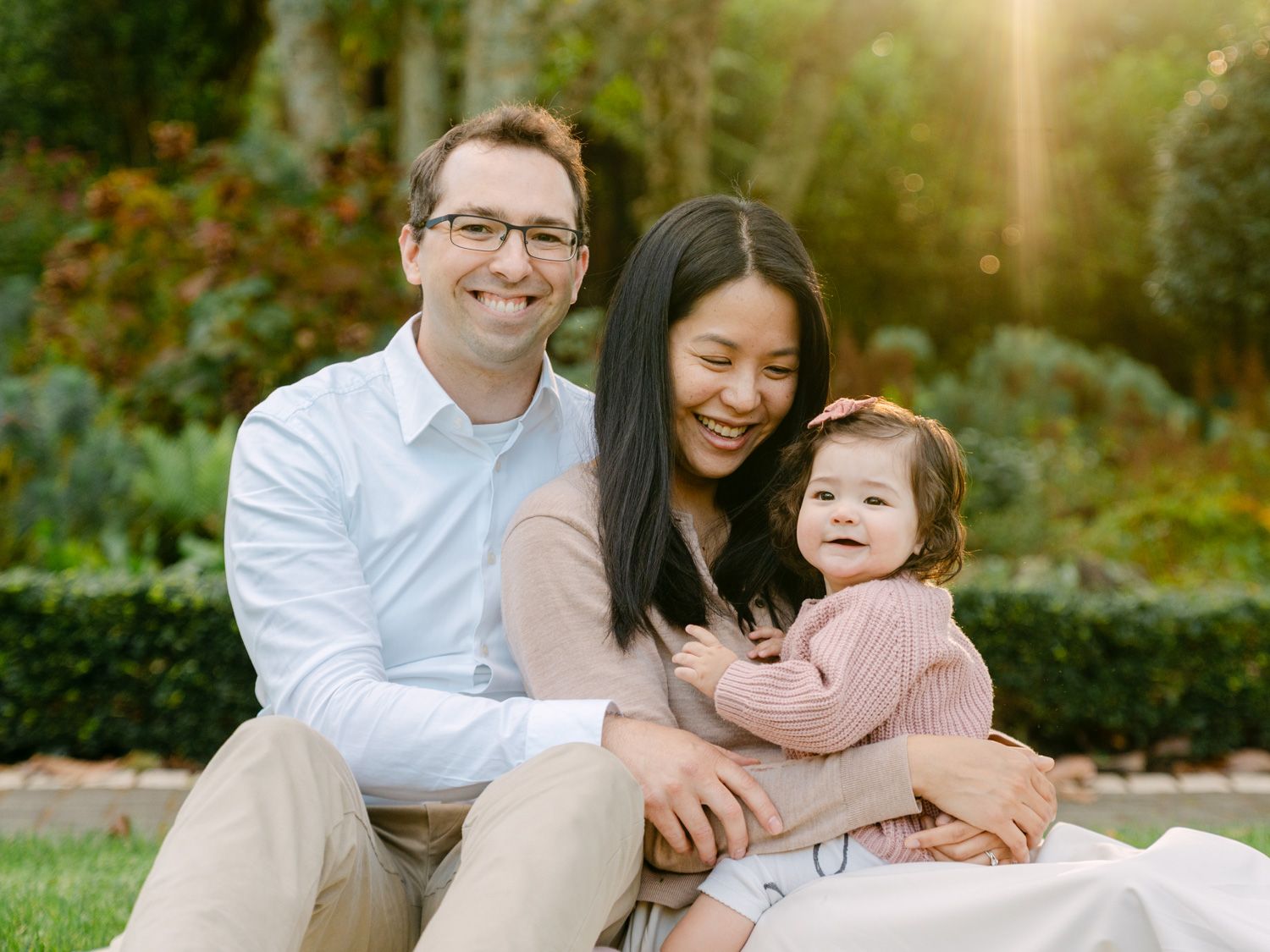 Happy family portrait with parents and baby girl sitting on grass in a sunlit garden setting.