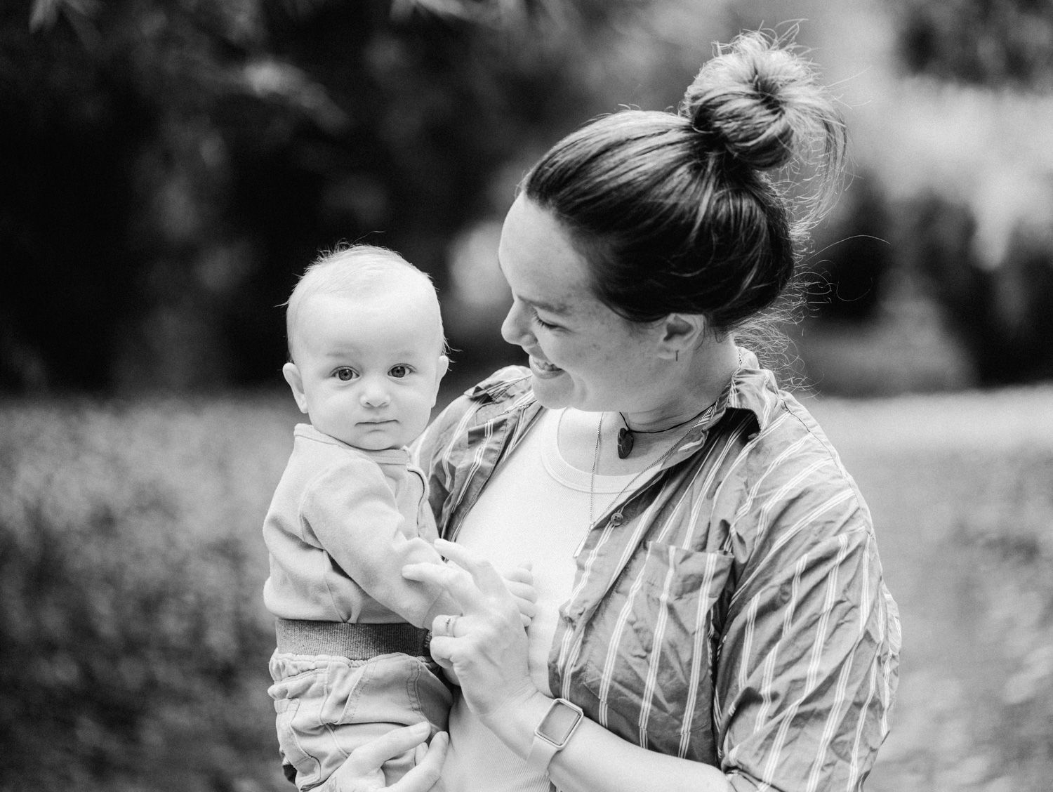Black and white photo of a smiling mother holding her baby outdoors in a garden setting.