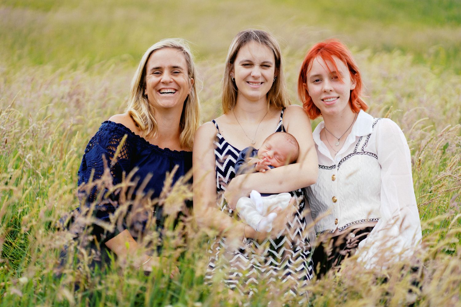 Three women sitting in a golden field, with one holding a newborn baby, smiling outdoors.