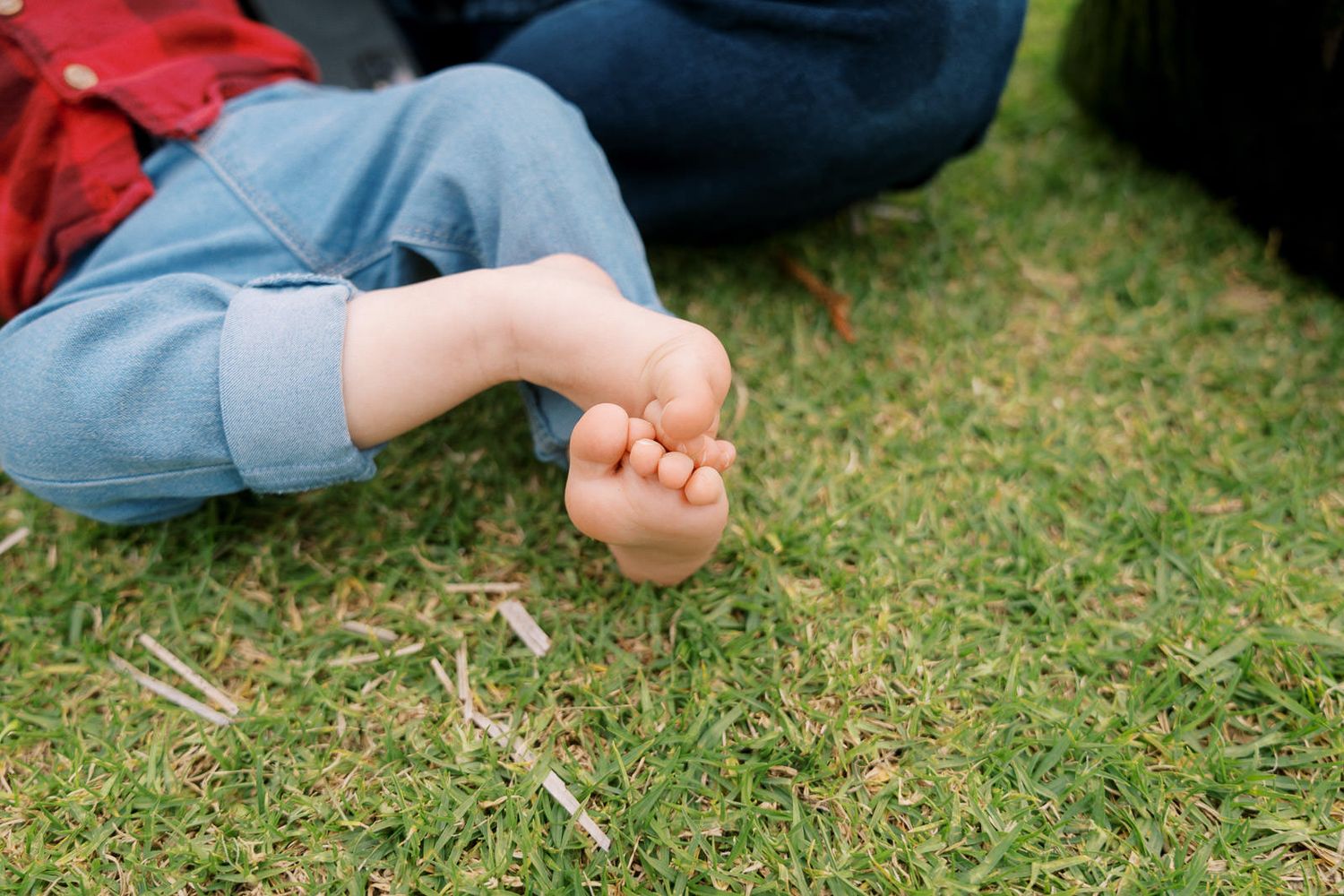 Bare baby feet resting on green grass outdoors, with child wearing blue denim jeans and red shirt.