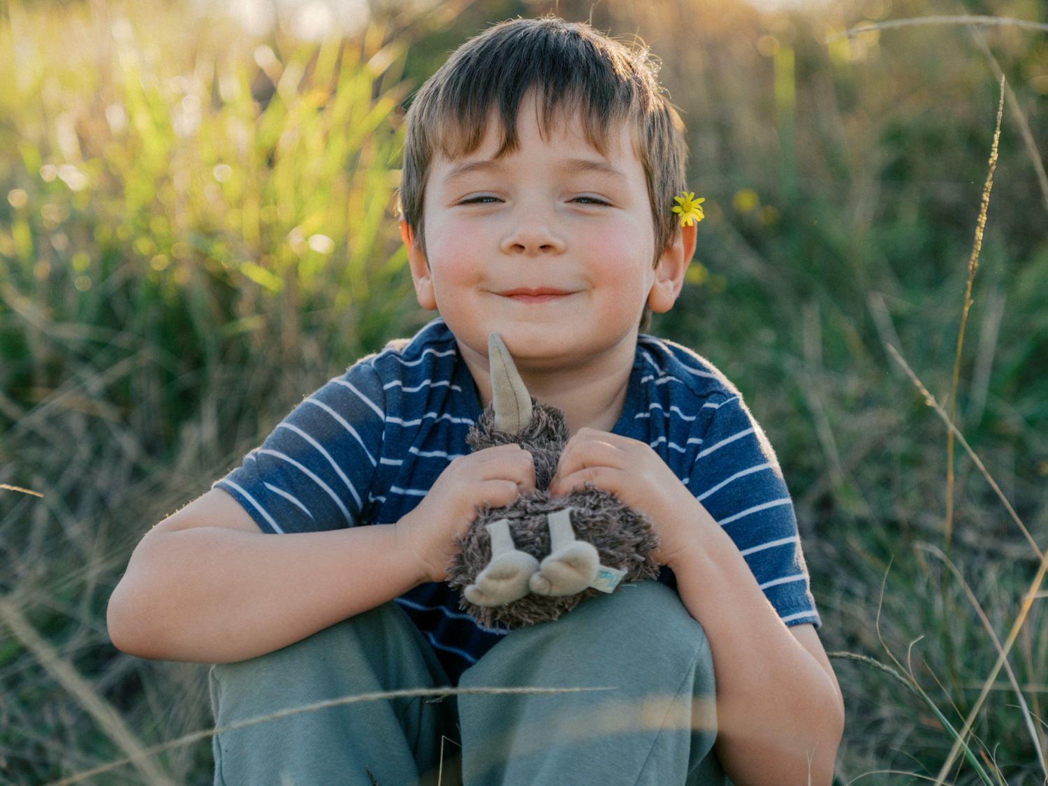 Smiling boy sitting in a sunny field holding a small rabbit, with a yellow flower tucked behind his ear.