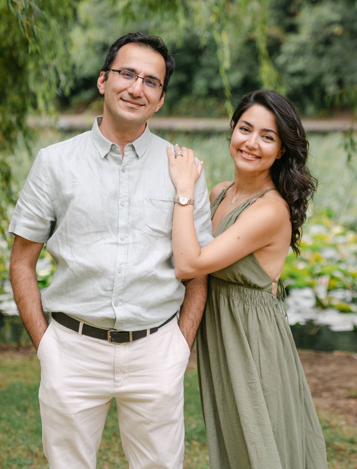 A smiling couple poses outdoors near a pond, woman in green dress with watch, man in gray shirt and white pants.