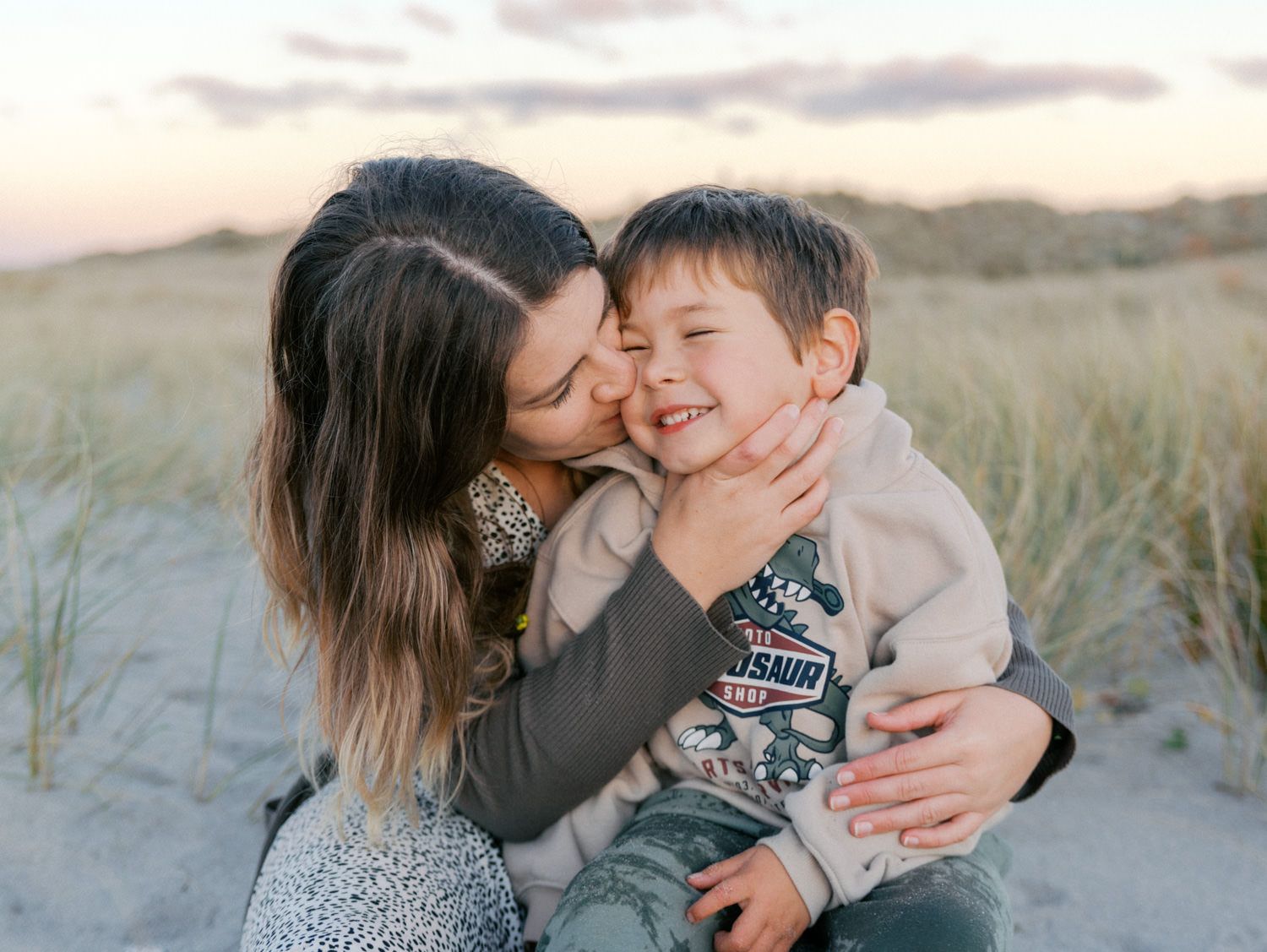 Mother kissing smiling young boy on cheek at sandy beach dunes during golden hour sunset.