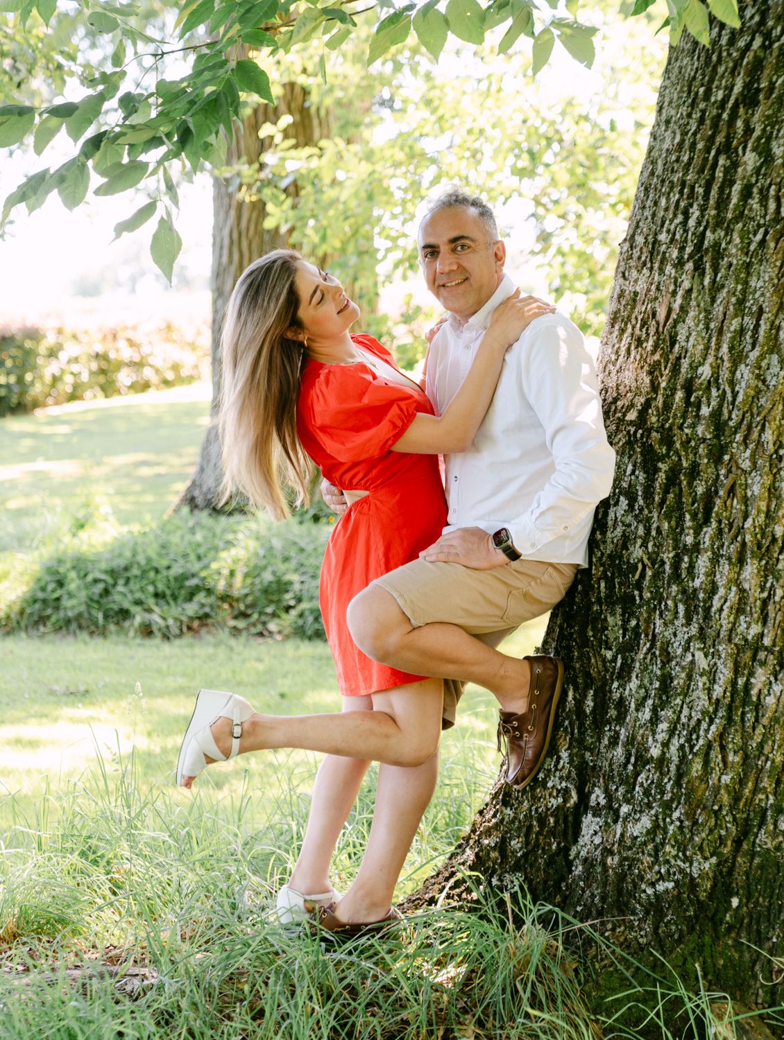 Couple embracing and laughing by a tree in a lush green park, woman in red dress, man in white shirt.
