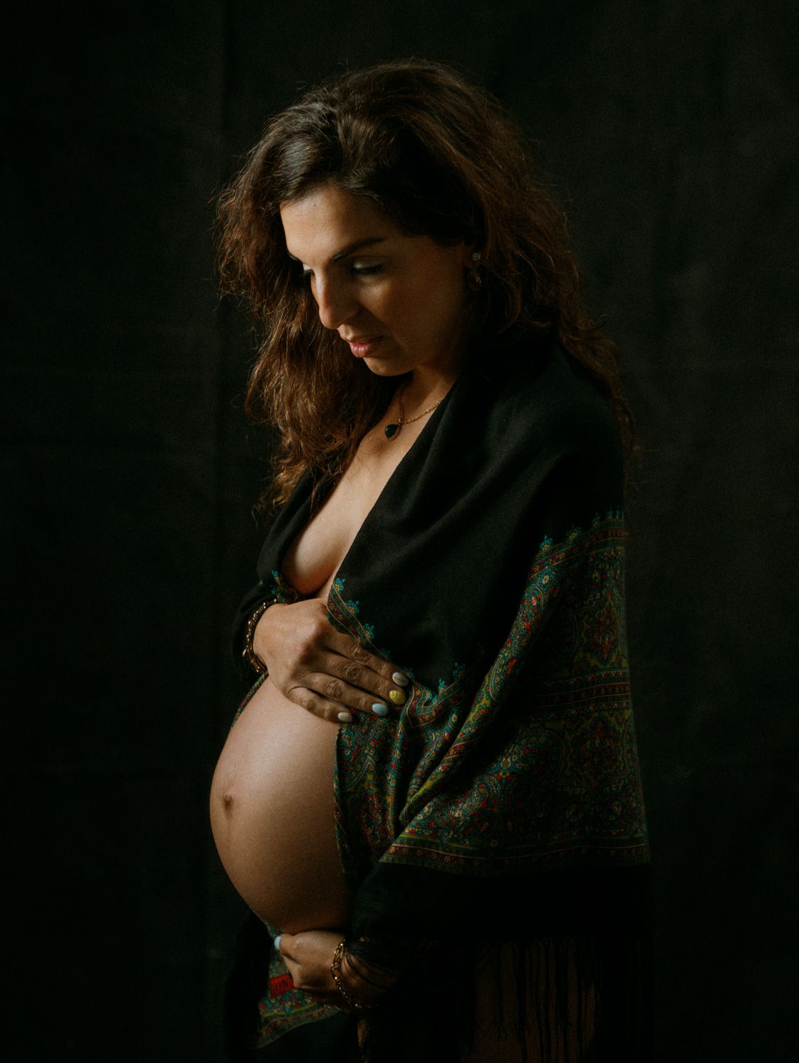 Pregnant woman in dark green floral wrap holding her baby bump in a moody studio portrait.