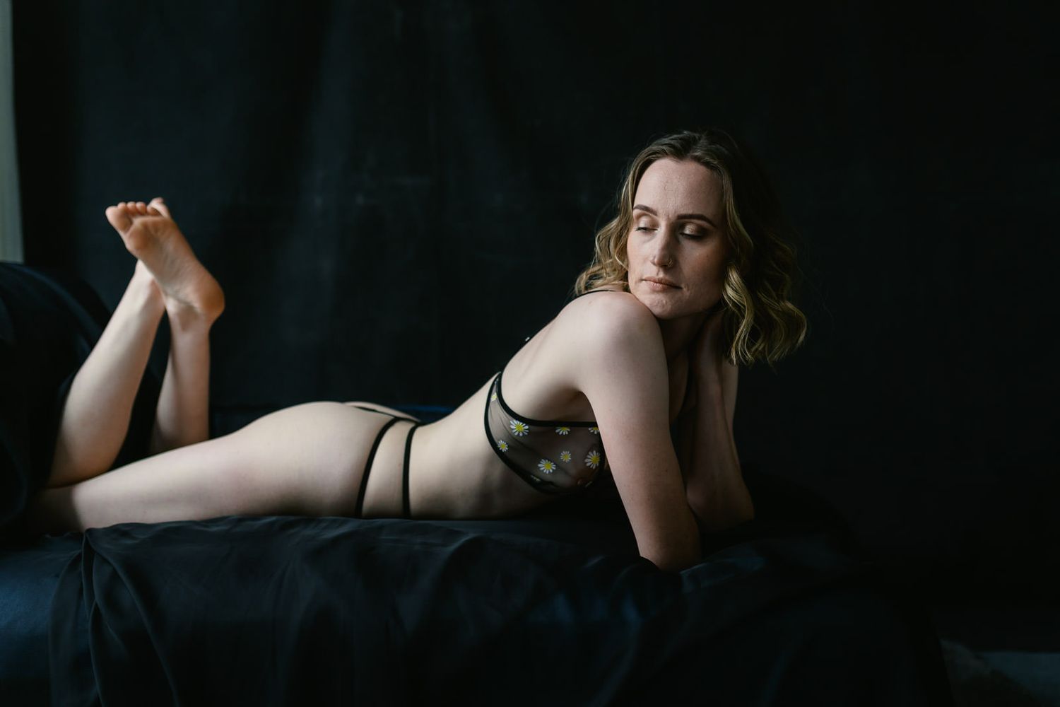 Woman in black lingerie posing on dark bedding against a black backdrop in a boudoir photography setting.