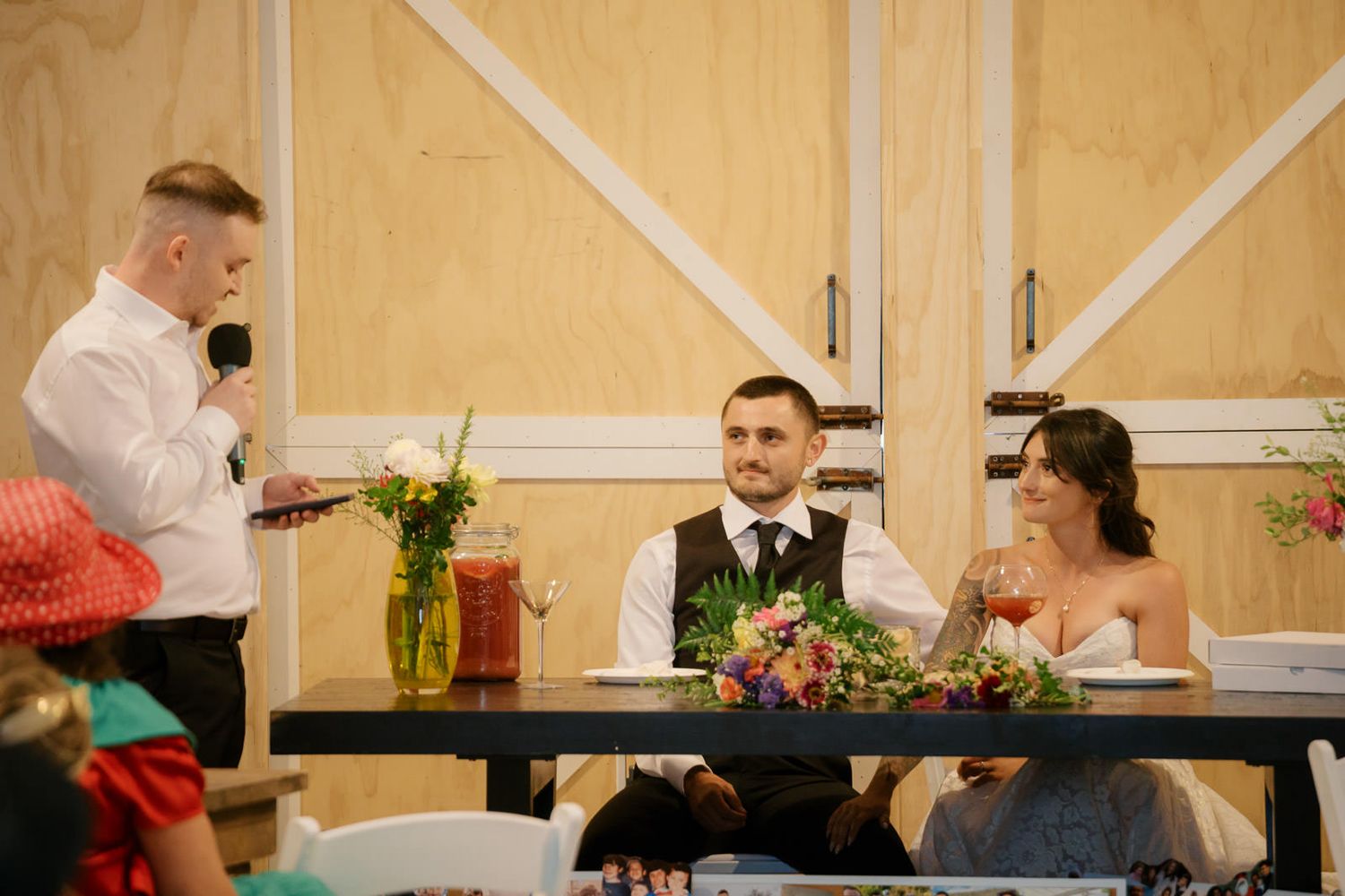 Best man delivers a heartfelt wedding speech to bride and groom seated at reception table in rustic barn venue.