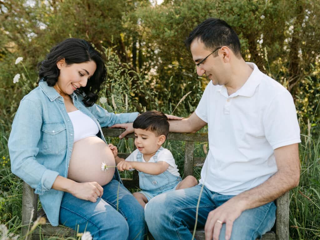 Pregnant woman and husband sit on bench outdoors with toddler son touching baby bump during family maternity photo session.