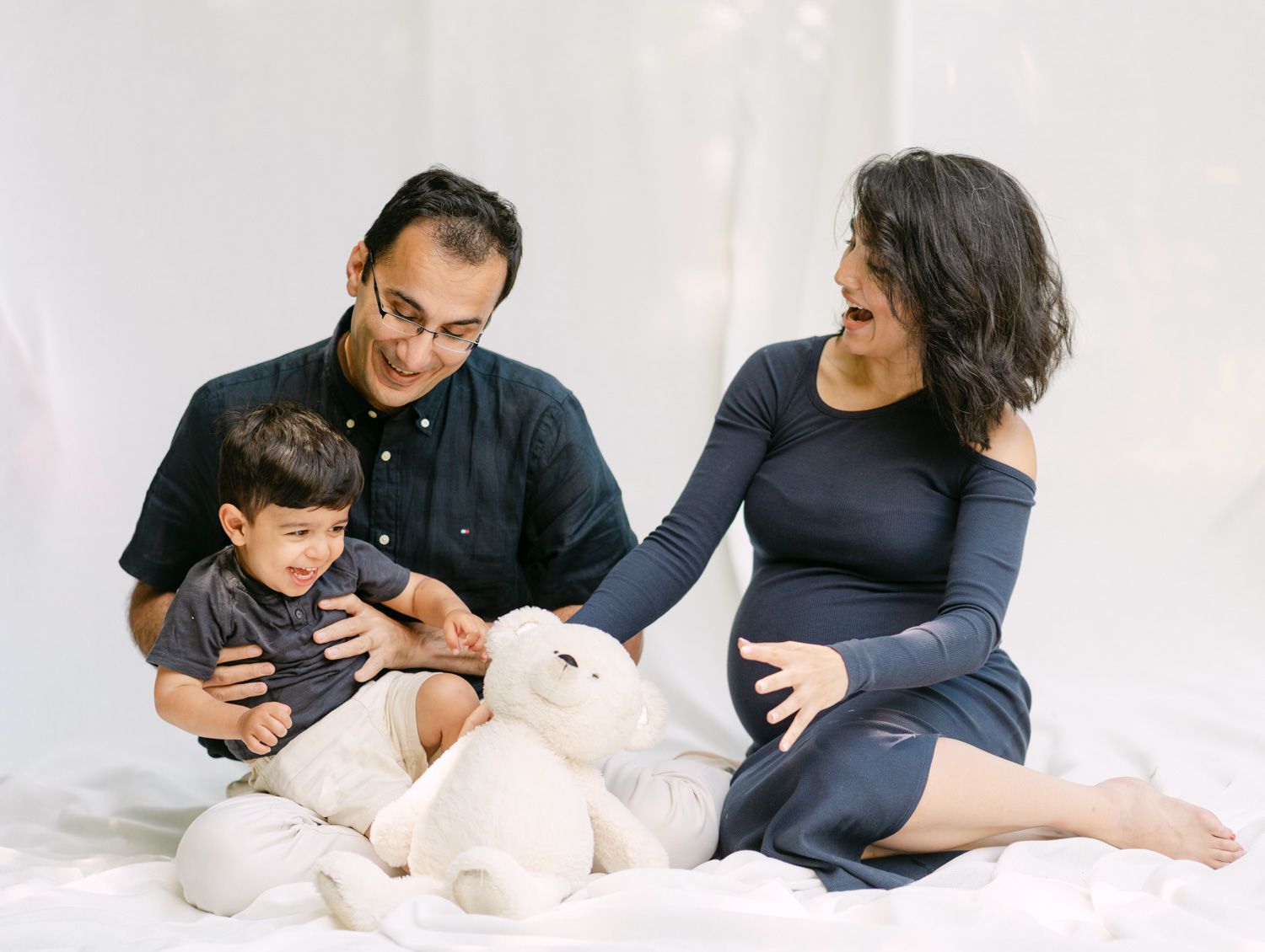 Pregnant woman in navy dress sits with husband and toddler son, posing with stuffed animal on white backdrop.