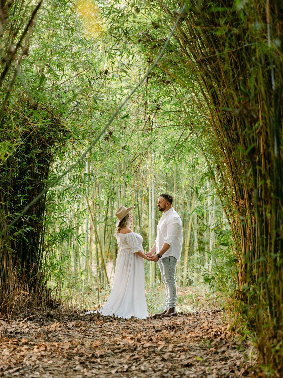 Couple holding hands in a lush bamboo forest, woman in white dress and hat, man in white shirt.