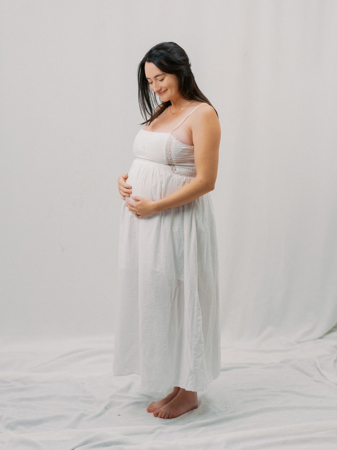 Pregnant woman in white maxi dress cradling baby bump, smiling softly in a bright studio setting.