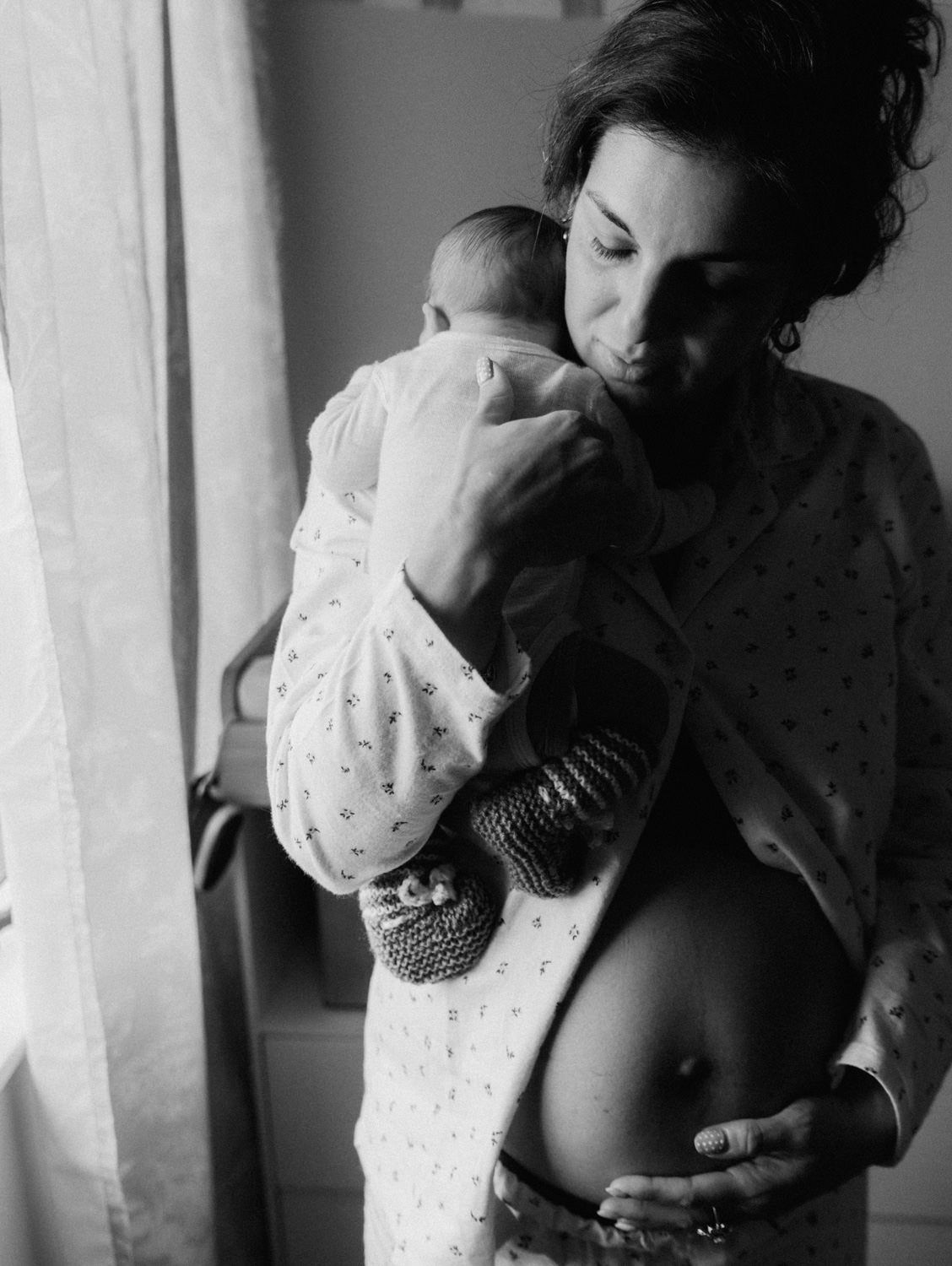 Black and white photo of a new mother in hospital gown holding her newborn baby while cradling her postpartum belly.