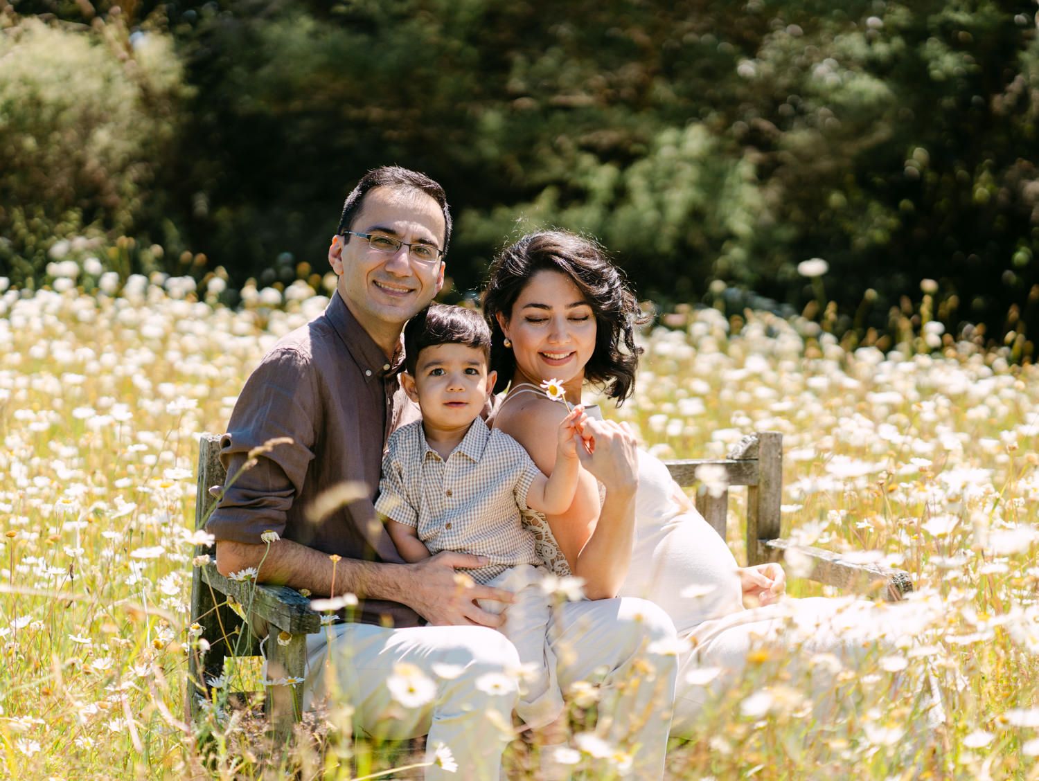 Happy family of three posing in a sunny daisy field, parents smiling with toddler on a wooden bench.