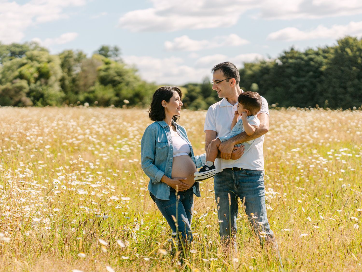 Pregnant woman and man holding toddler stand in sunny wildflower field, family maternity photo outdoors.