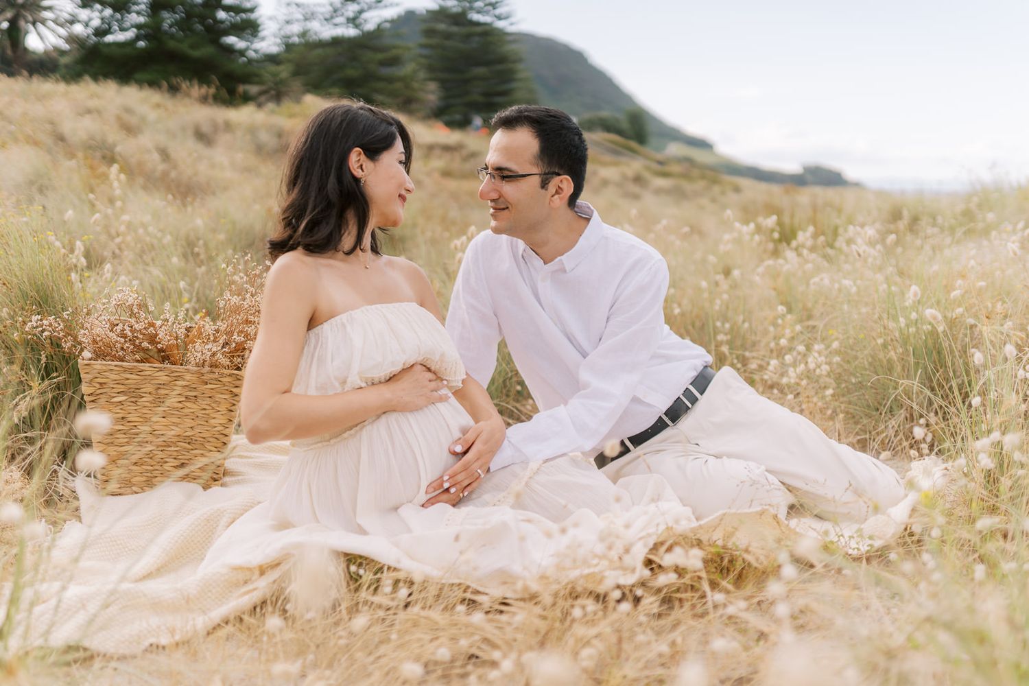Couple sitting in golden field for maternity photoshoot, woman in white dress cradling baby bump, man in white outfit beside her.