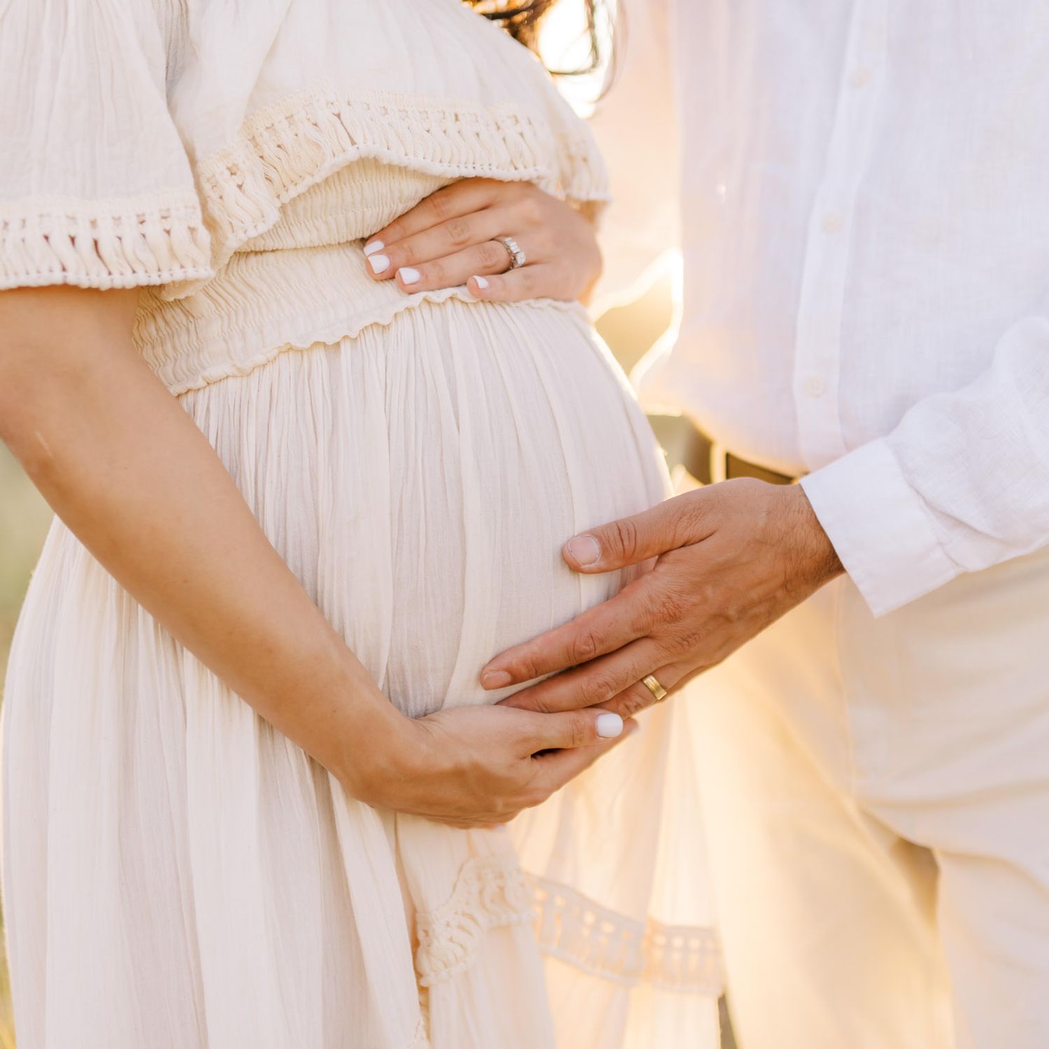 A pregnant woman in a white dress with her partner both cradling her baby bump outdoors.