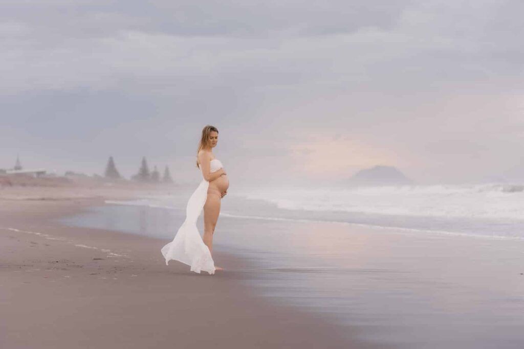 A pregnant woman in a flowing white dress poses gracefully on a serene beach at sunset.