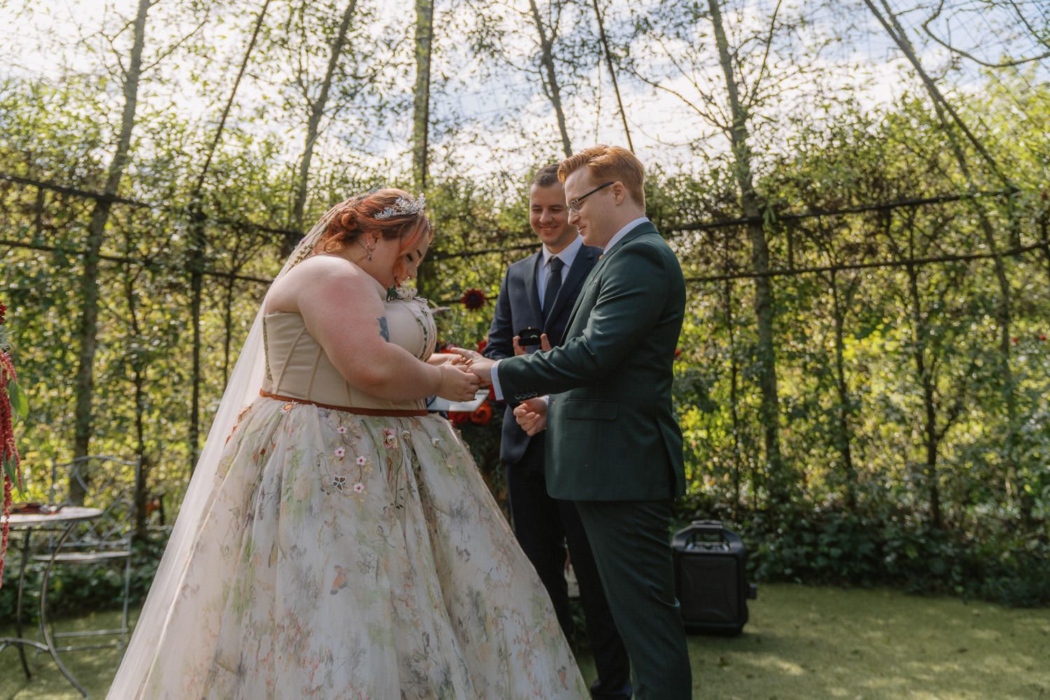 Couple exchanging rings during outdoor wedding ceremony in lush garden gazebo, bride in floral gown, groom in green suit.