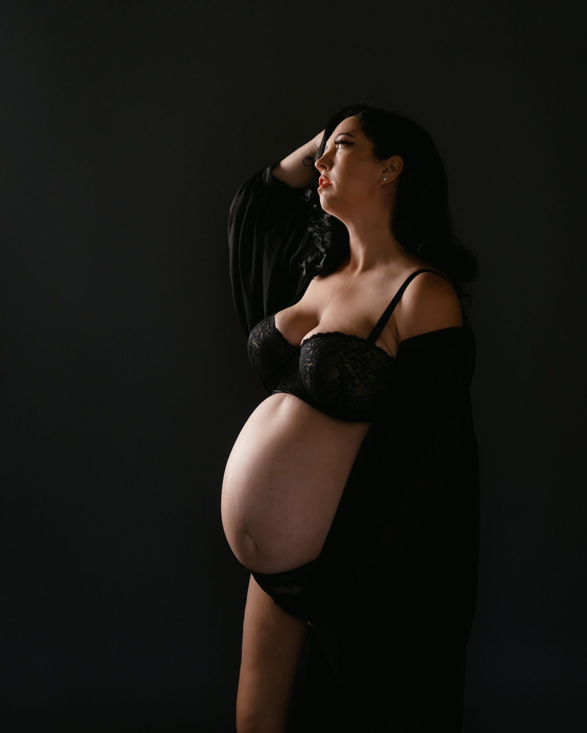 Pregnant woman in black lace lingerie posing against dark background in artistic maternity portrait photography.