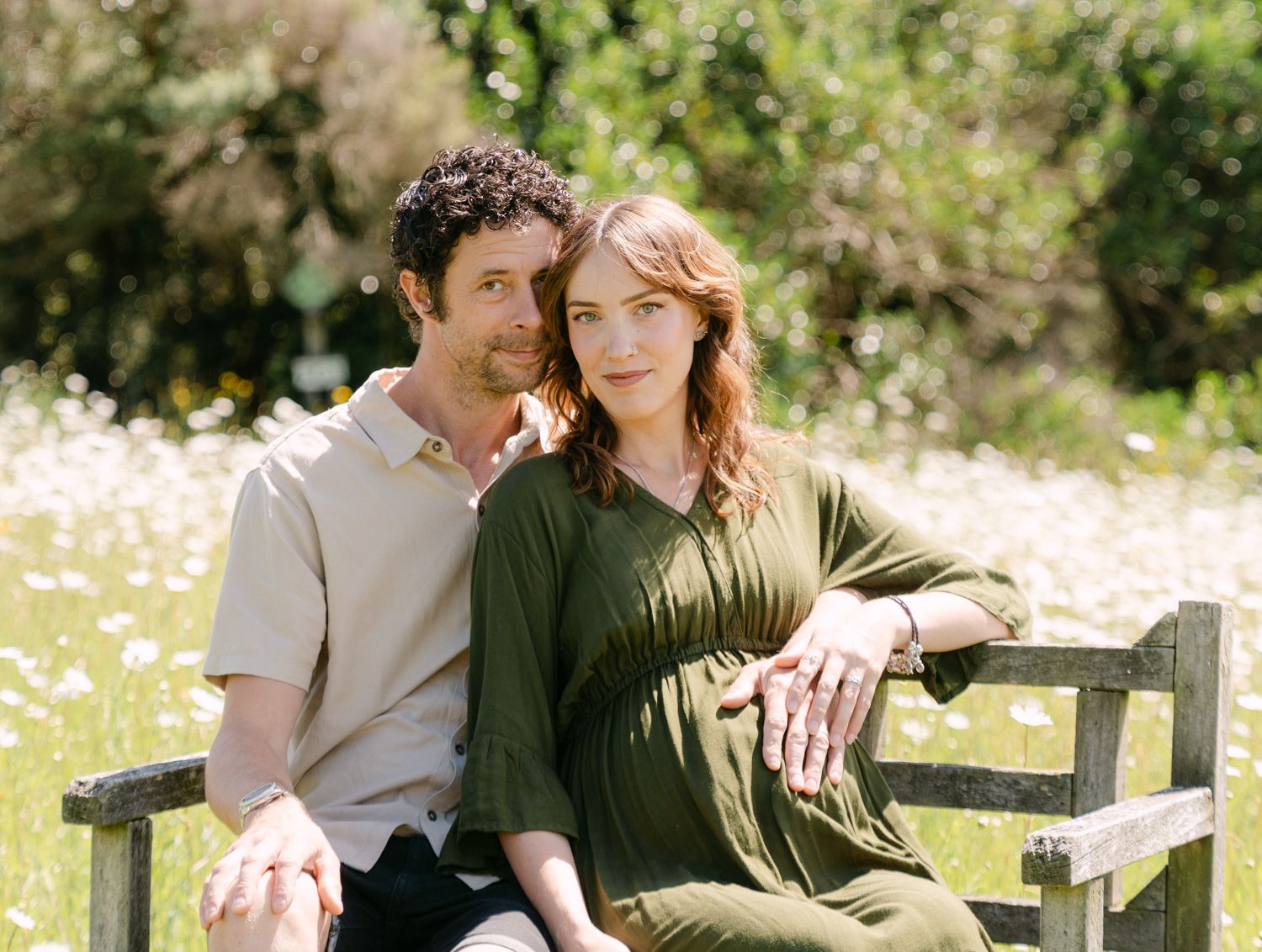 Couple sitting together on a wooden bench in a sunny garden with wildflowers, woman in green dress, man in beige shirt.