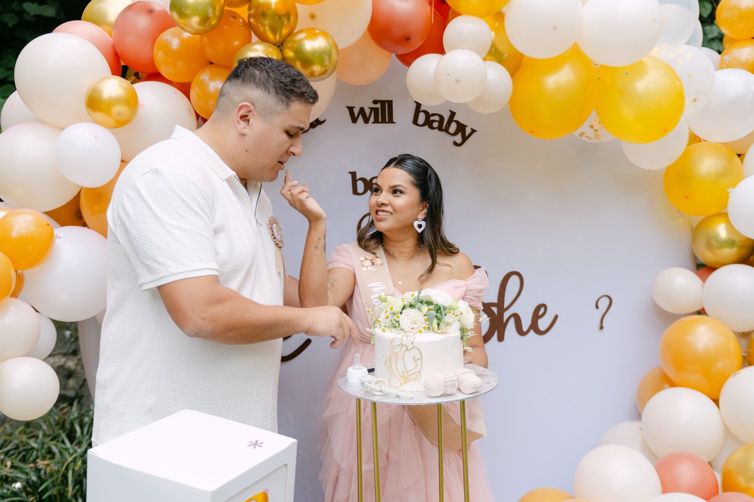 Couple at gender reveal party with balloon arch and cake, sign reads will baby be a he or she.