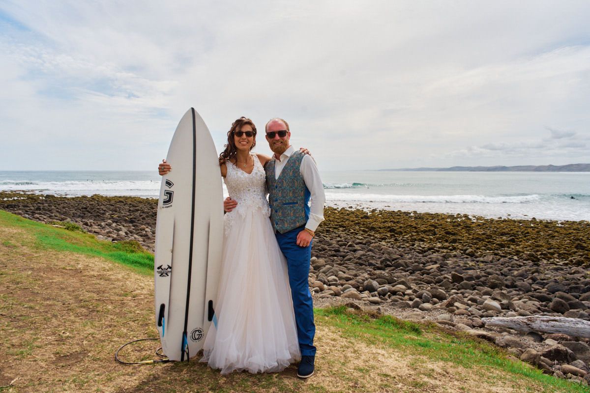 Bride and groom posing with a white surfboard near rocky shoreline on their beach wedding day.