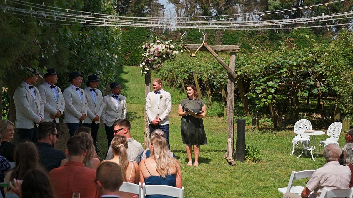 Outdoor wedding ceremony in a lush garden with guests seated, groom at wooden arch, officiant, and groomsmen standing nearby.