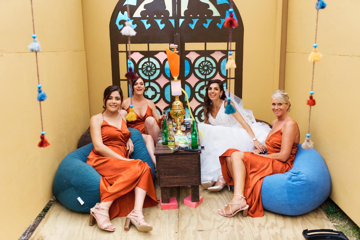 Bride in white dress with three bridesmaids in orange gowns relaxing on bean bag chairs at a colorful wedding celebration.