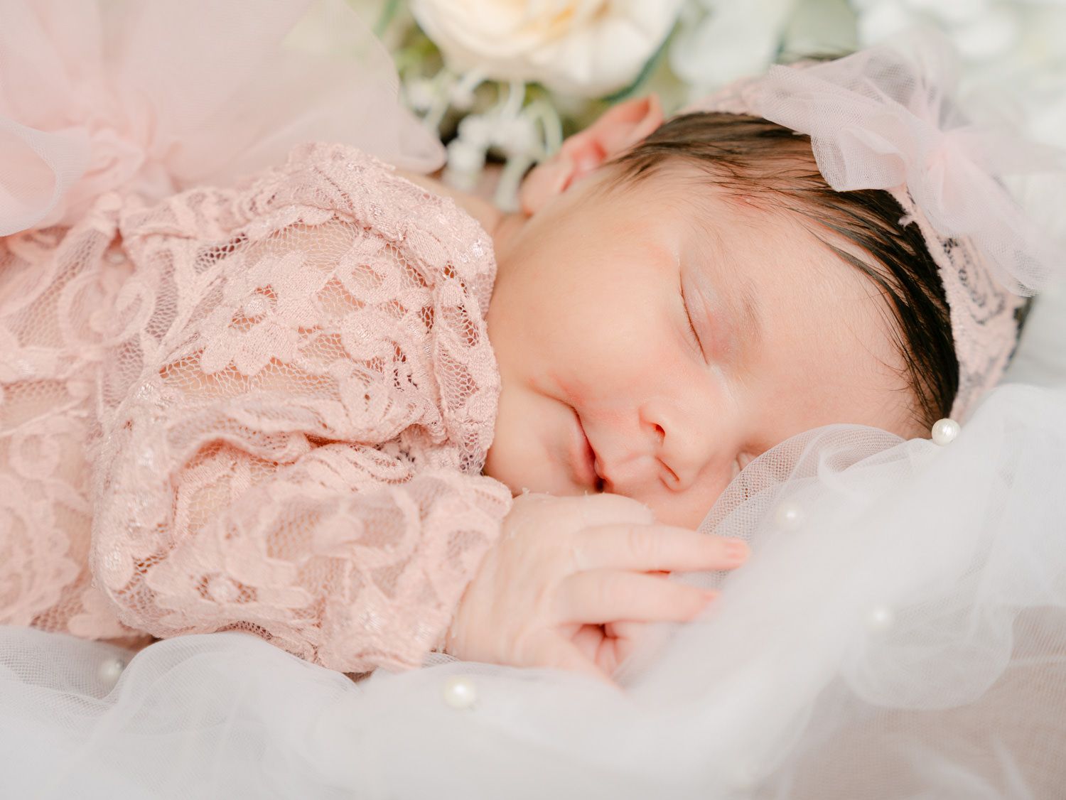 Sleeping newborn baby in pink lace outfit and floral headband, peacefully resting on white tulle.