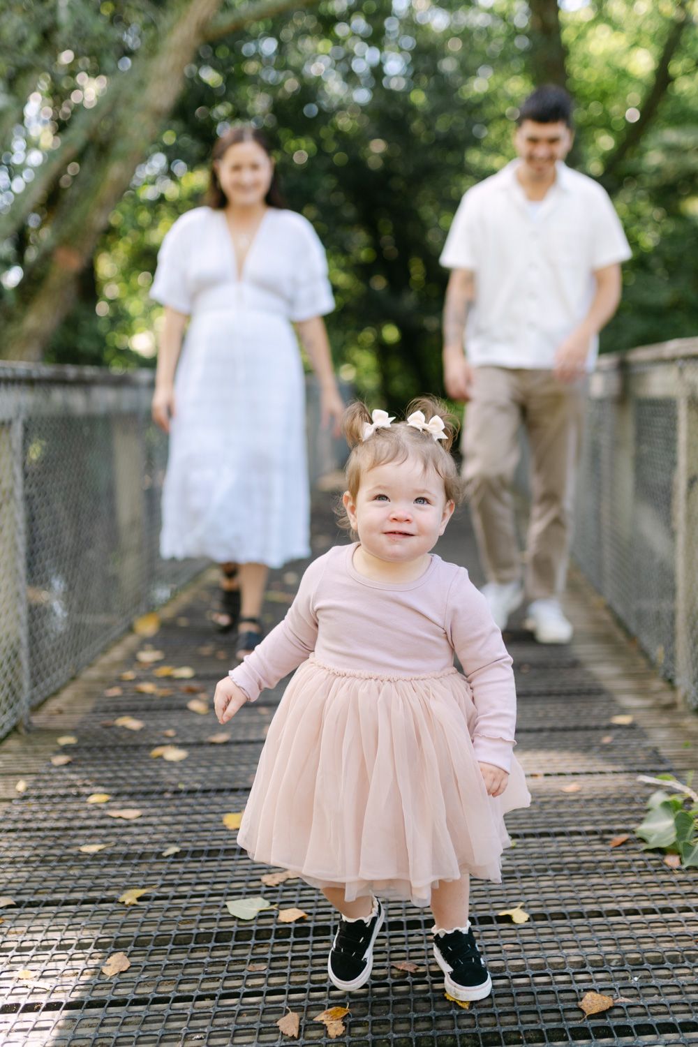 Toddler girl in pink dress on bridge with parents in white outfits standing behind her in a green park.
