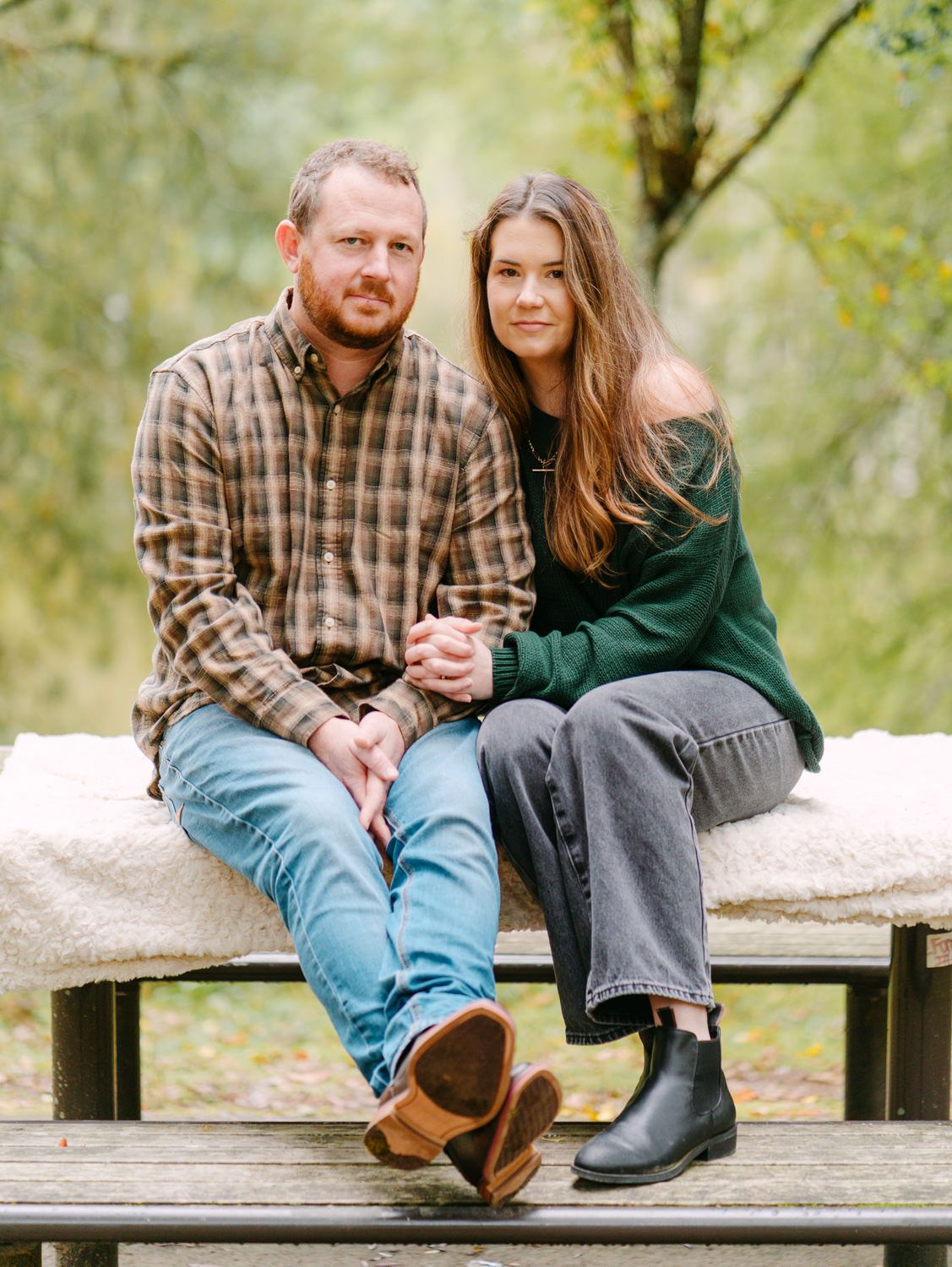 A couple sits together on a picnic table bench outdoors, surrounded by autumn foliage, smiling warmly.