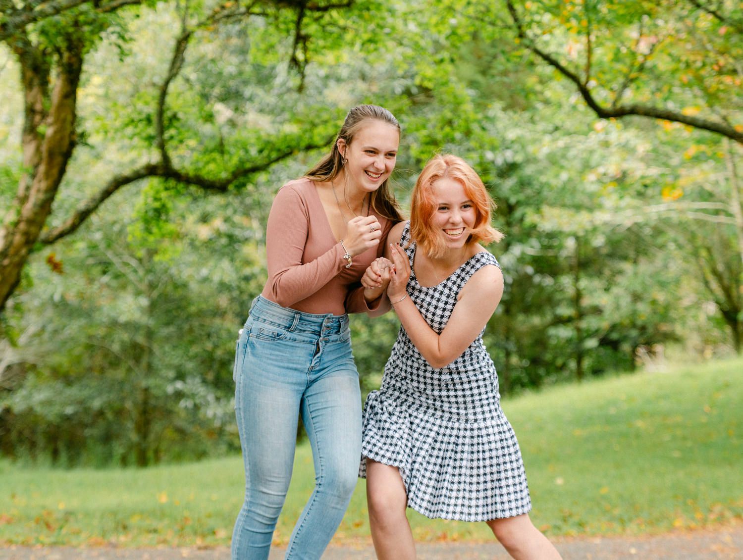 Two young women laughing and posing playfully in a lush green park, one in jeans and one in a gingham dress.