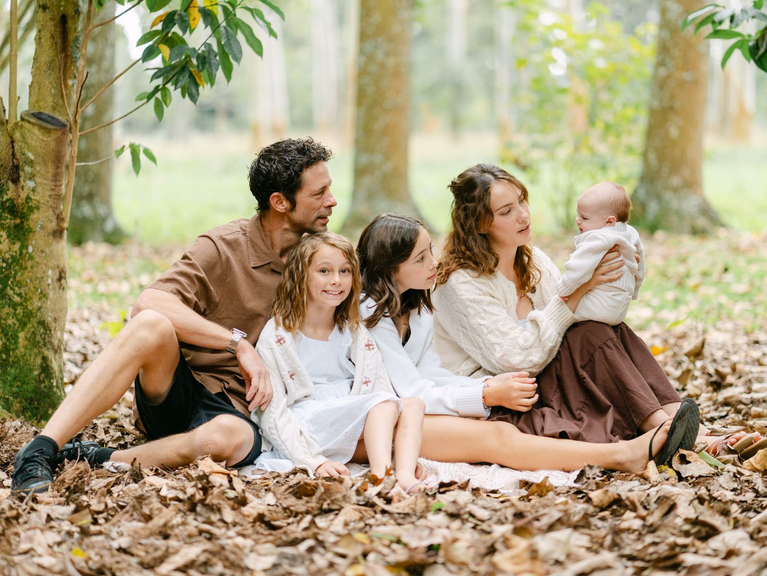 Family of five sitting together on autumn leaves in a forest, sharing a warm and joyful outdoor moment.