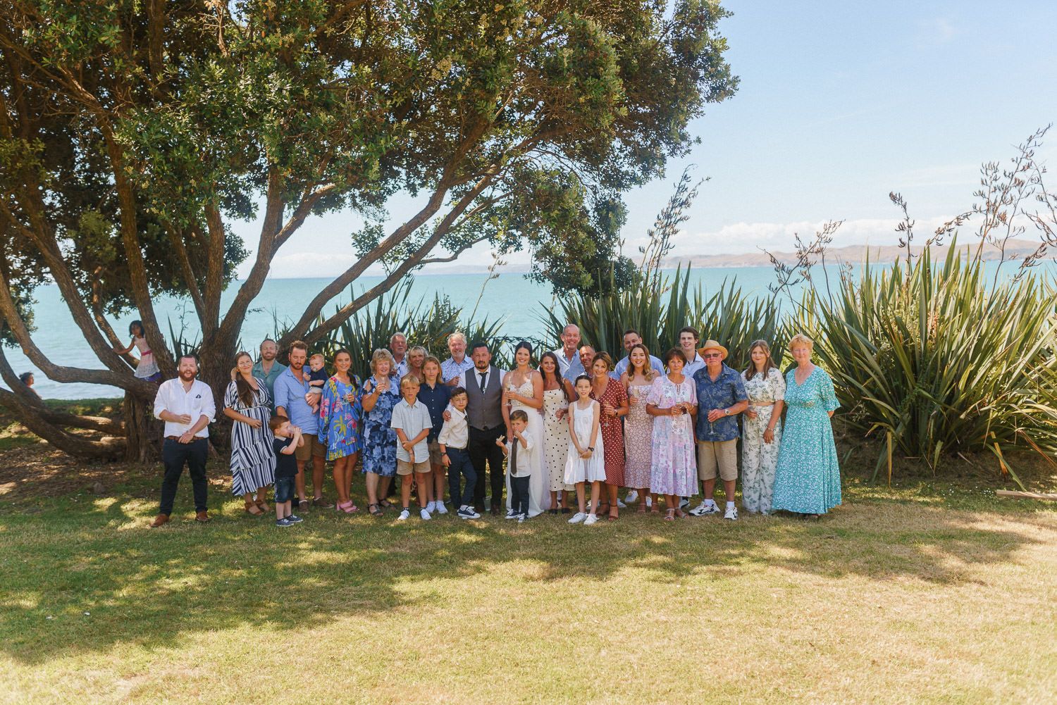 Large group of people posing outdoors near trees with ocean views on a sunny day.