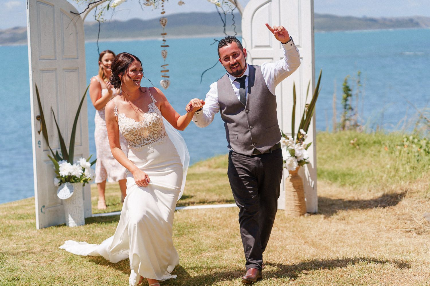 Joyful newlyweds celebrate after outdoor waterfront wedding ceremony, bride in white gown, groom in gray vest.