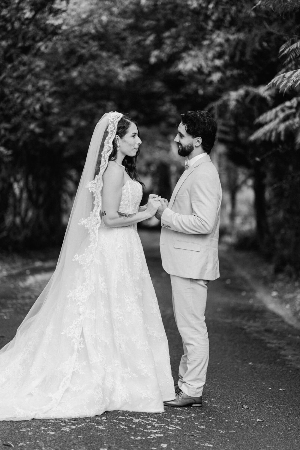 Black and white photo of bride in lace gown and veil holding hands with groom in light suit on tree-lined path.