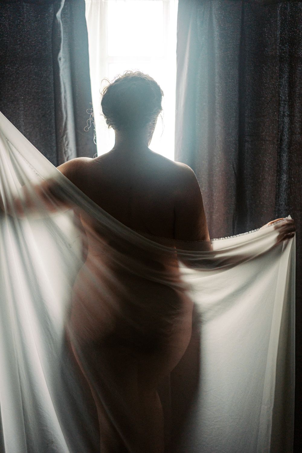Silhouette of a person holding sheer white curtains in front of a bright window with soft backlight.