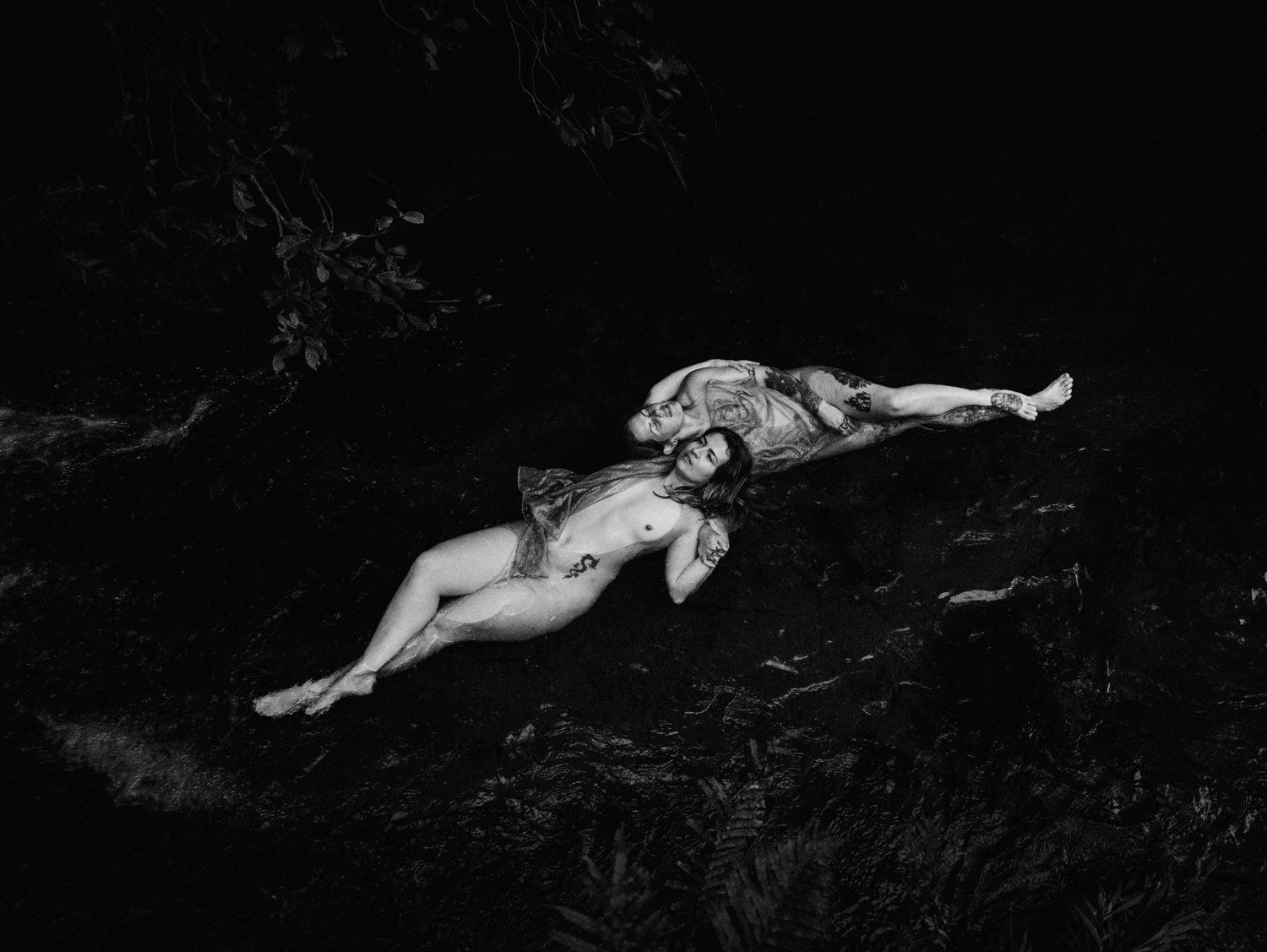 Black and white fine art photo of two nude figures reclining on dark rocks surrounded by lush ferns.
