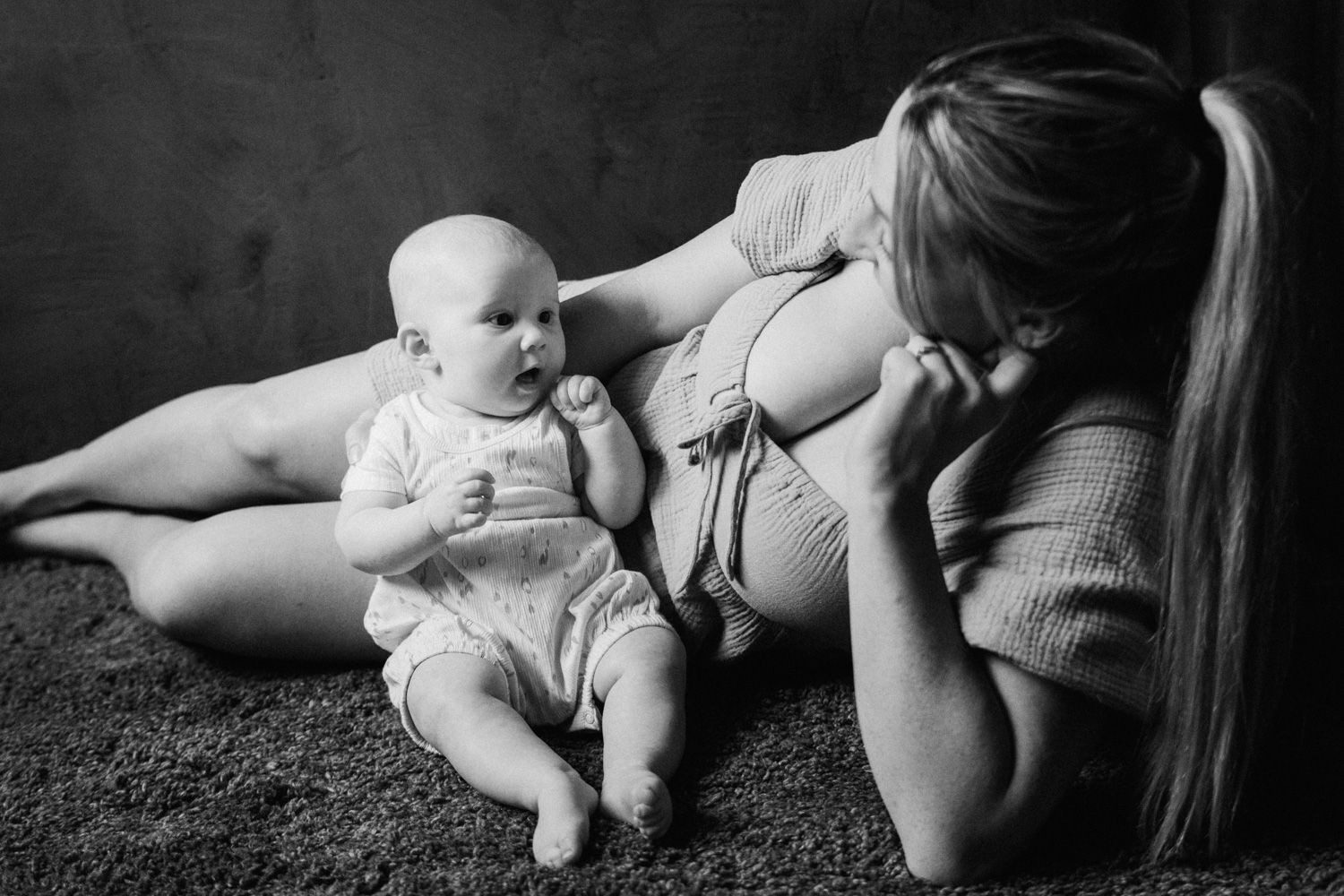 Black and white photo of a mother lying on carpet gazing lovingly at her baby sitting beside her.