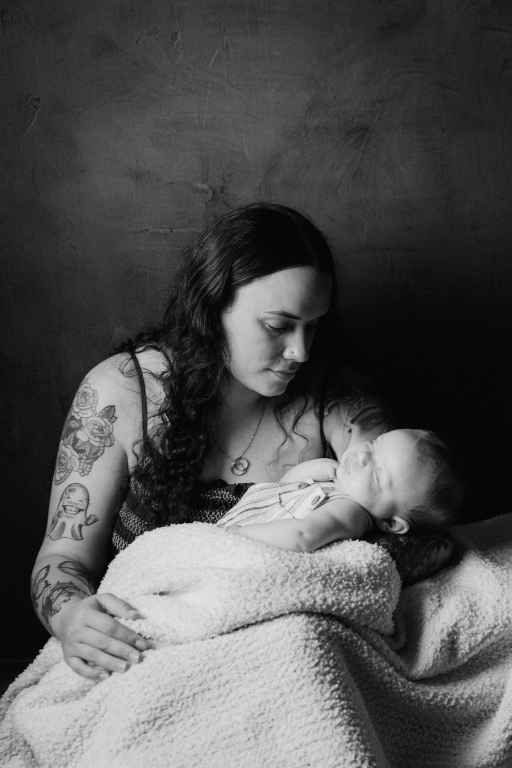 Black and white photo of tattooed mother tenderly holding sleeping newborn wrapped in white blanket.
