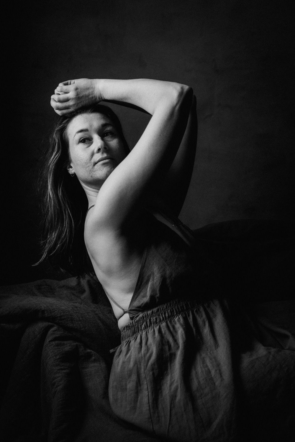 Black and white portrait of a woman in a linen dress, arms raised gracefully above her head against a dark background.