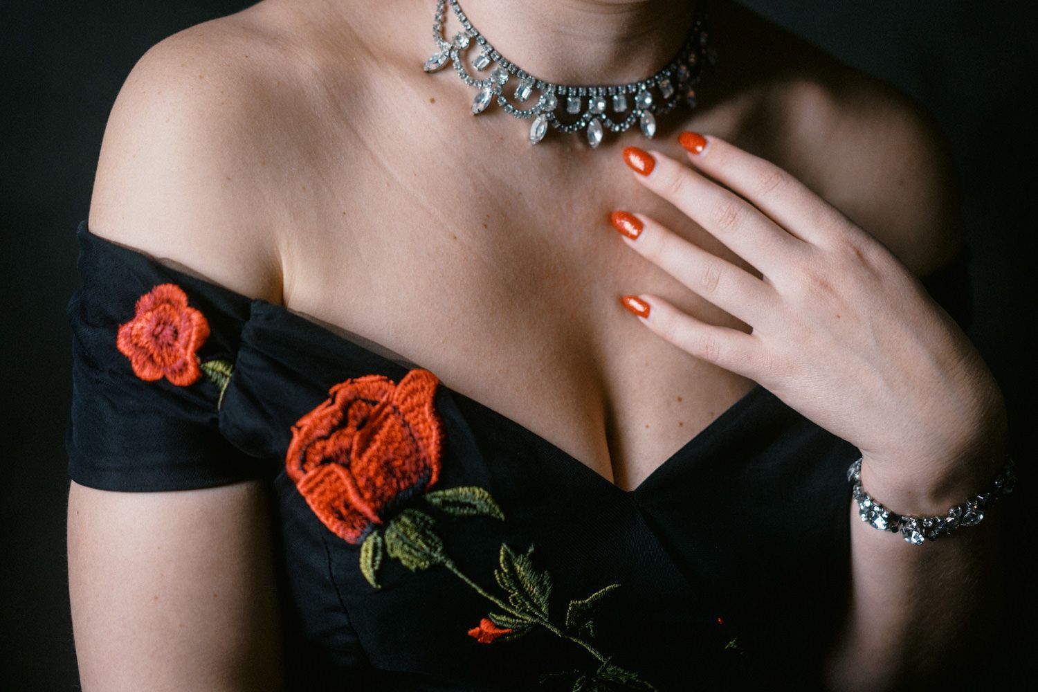 Woman in black off-shoulder dress with red rose embroidery, wearing diamond necklace and bracelet with orange nails.