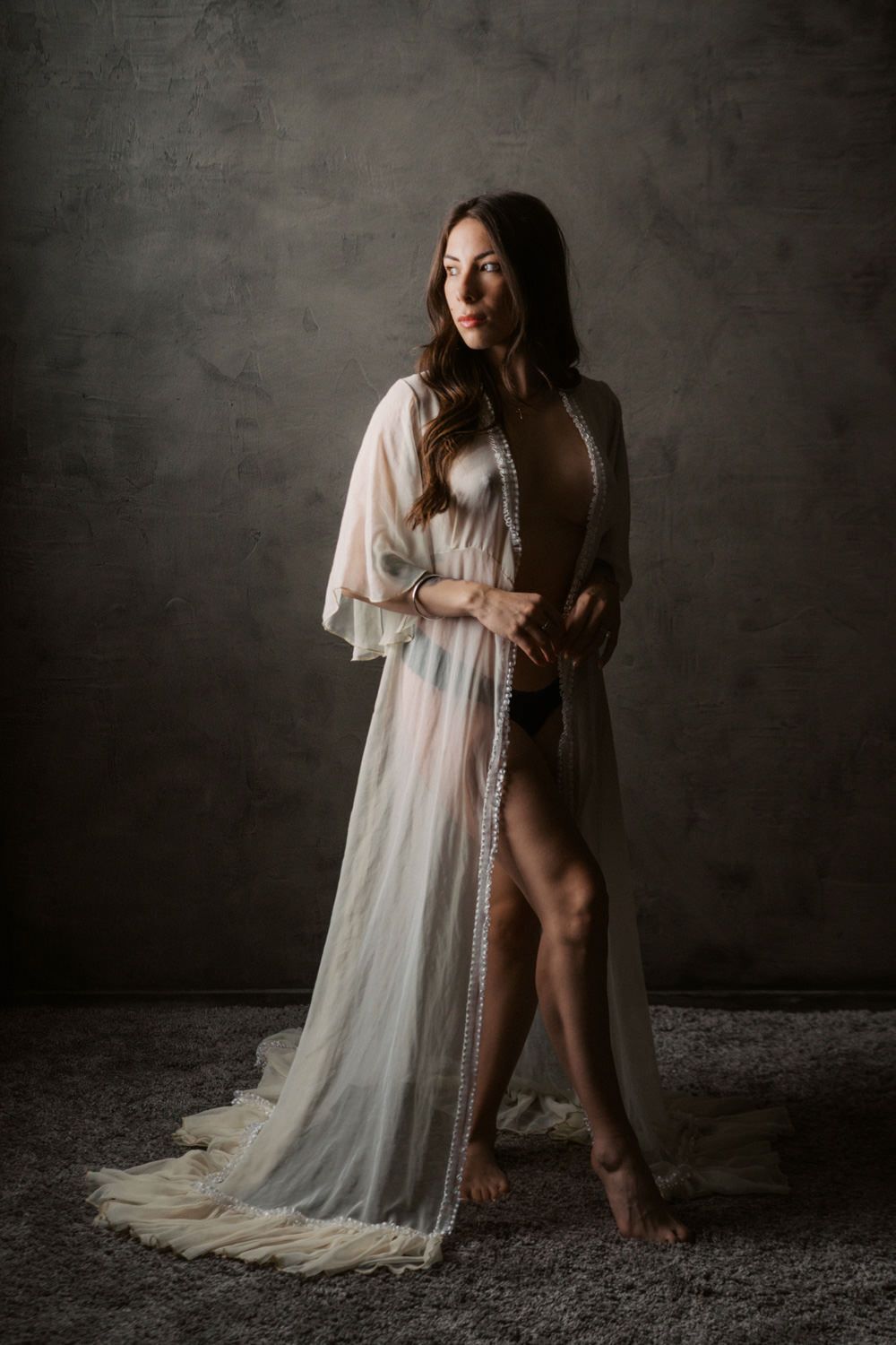 Woman in sheer white maxi robe posing elegantly in a studio with gray textured background.